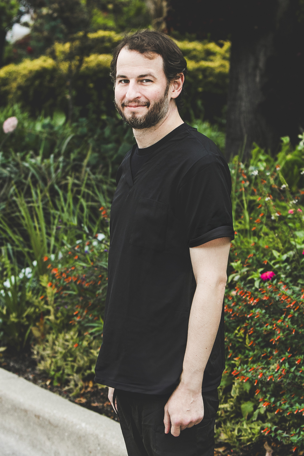 A man with a fair, complexion, short, wavy brown hair and brown beard with flecks of grey and wearing a black medical outfit is seen standing in front of an urban garden on a bright summer day