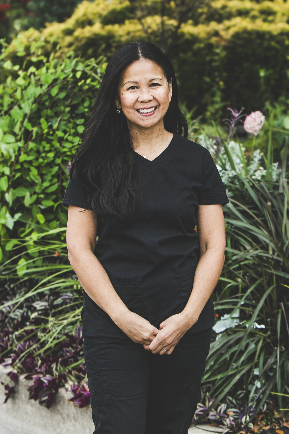 A woman with a medium complexion, long, dark straight black hair and wearing a black medical outfit is seen standing in front of an urban garden on a bright summer day