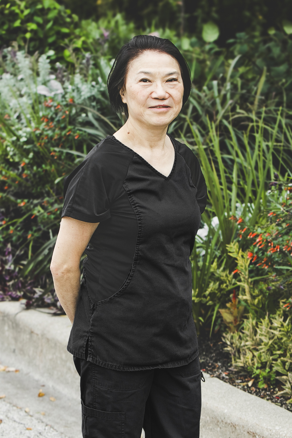 A woman with short, straight, neck-length black hair and wearing a black medical outfit is seen standing in front of an urban garden on a bright summer day