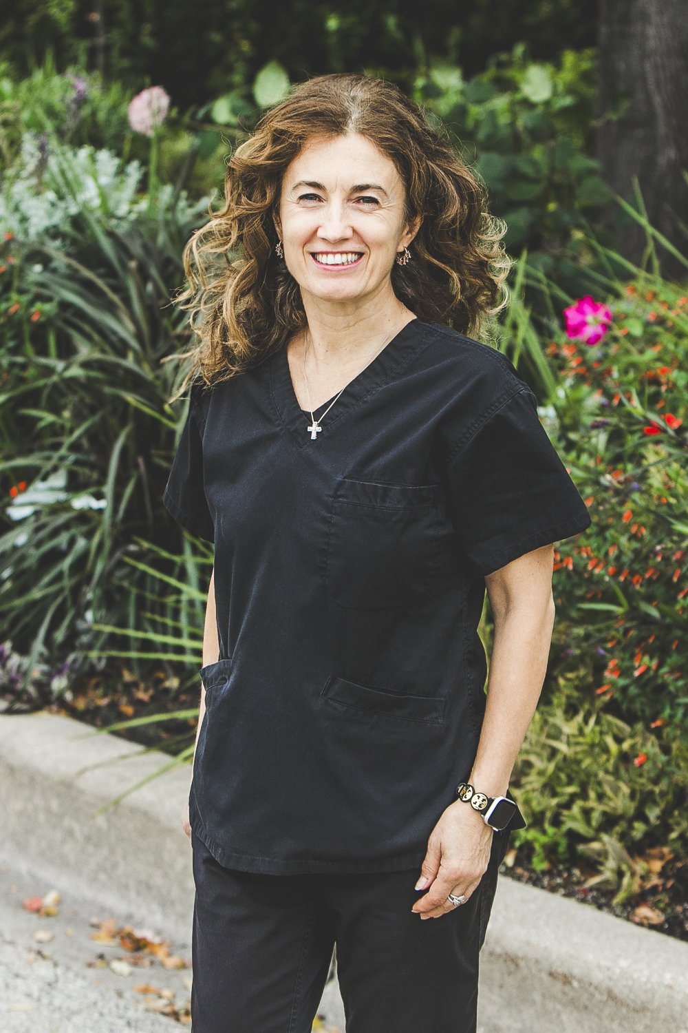 A woman with a fair complexion with slightly-curly shoulder-length brown hair with highlights wearing a dark-grey medical outfit is seen standing in front of an urban garden on a bright summer day