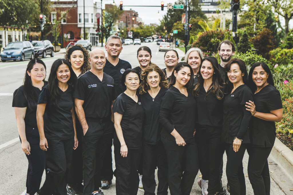 A group of dentists and dental technicians in dark-colored medical outfits are standing on a city street mid-day and smiling.