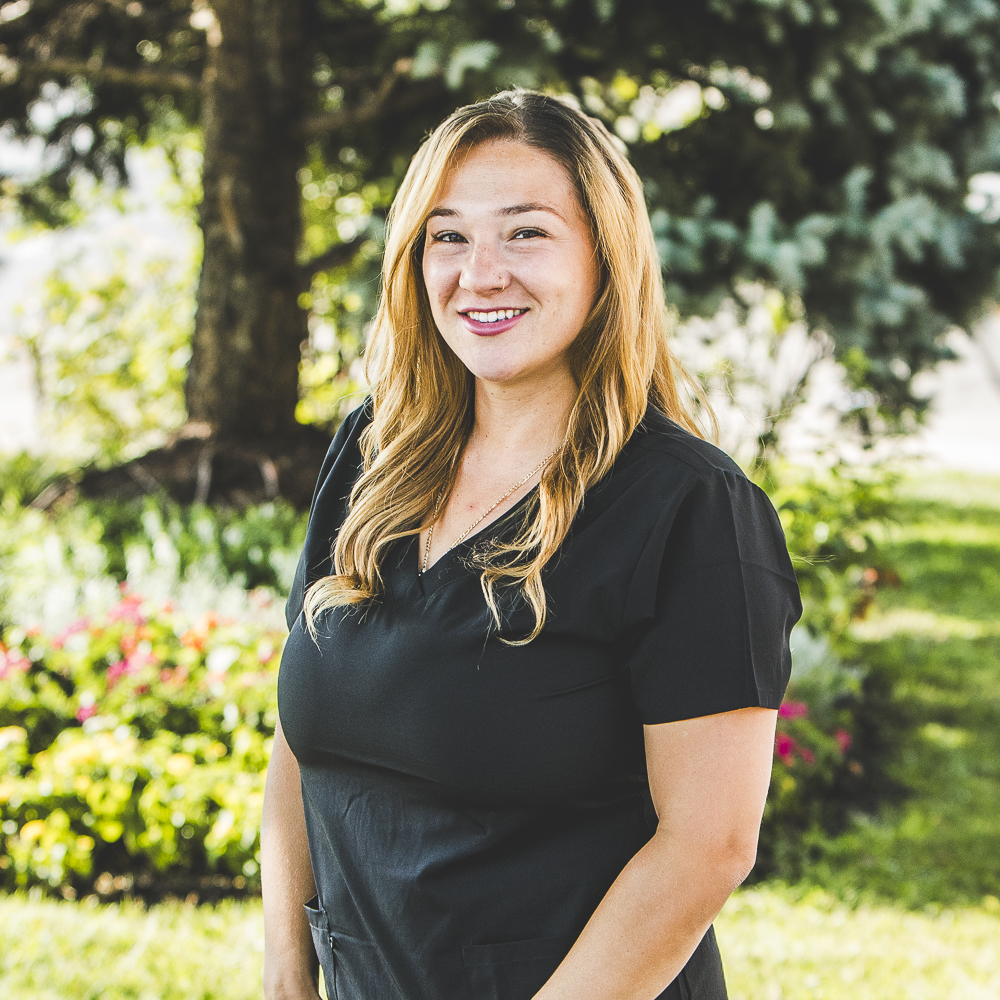 A woman with a tan complexion and long, flowing, sandy blonde hair, and wearing a black medical outfit, is seen standing in front of a garden on during sunny daytime weather